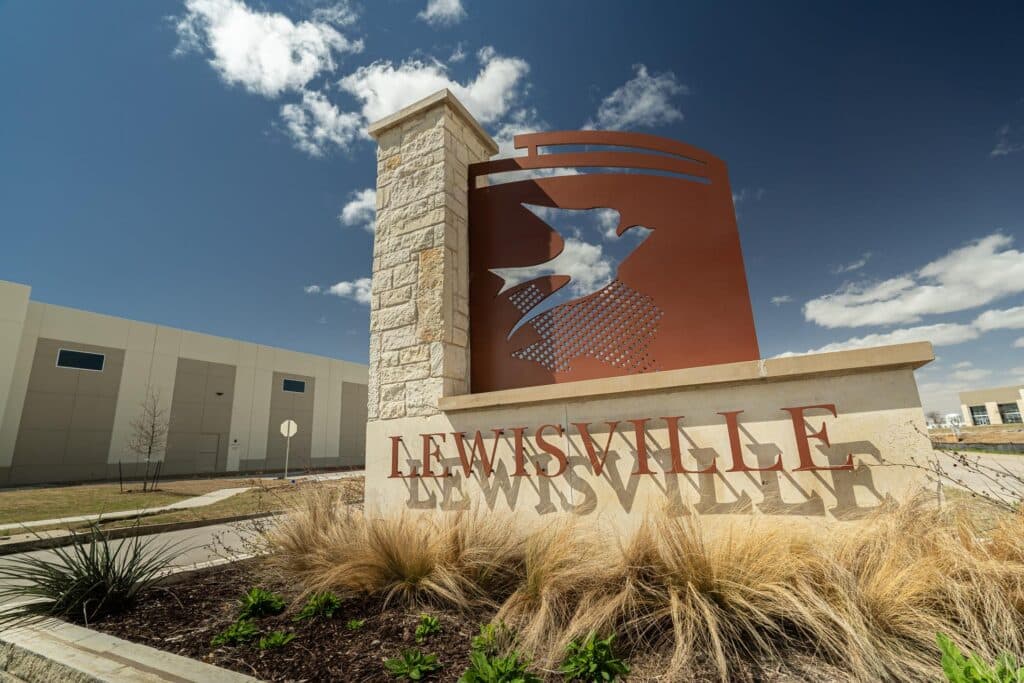 Lewisville city sign with a metal sculpture and stone pillar under a blue sky.